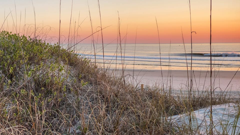 A beach at sunset with waves, sand dunes, and sparse vegetation.