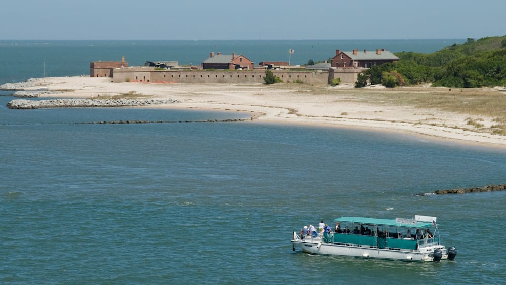A boat on calm waters near a sandy beach with a historic building in the background.