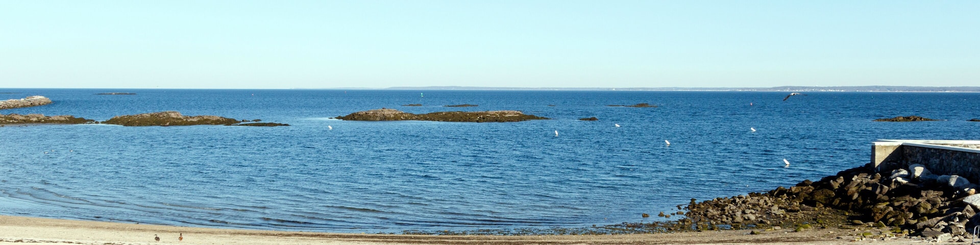 Un paisaje costero con una playa arenosa, aguas azules tranquilas y afloramientos rocosos.