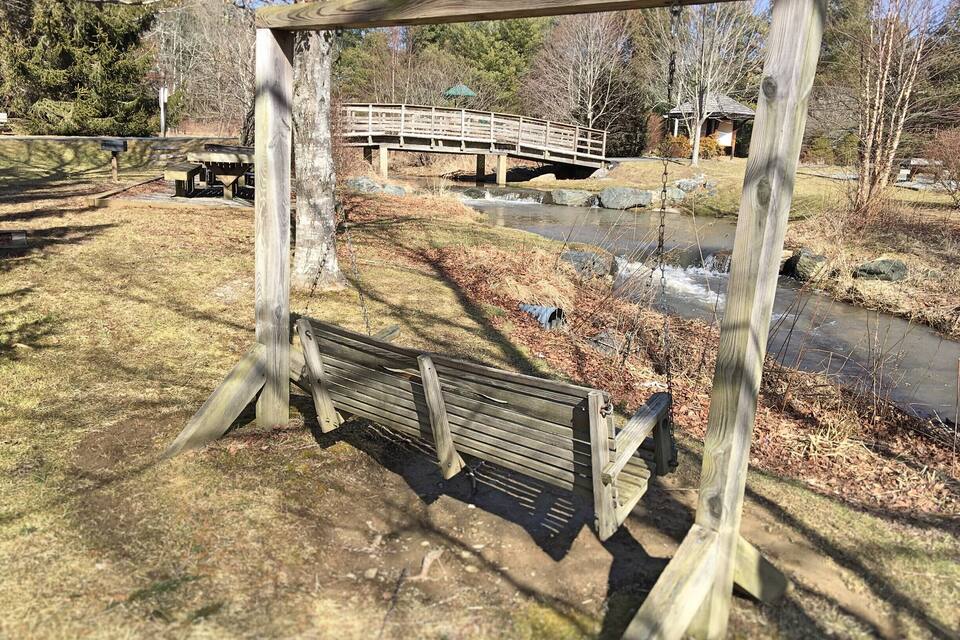 A wooden swing set in a park with a small stream and a wooden bridge in the background.