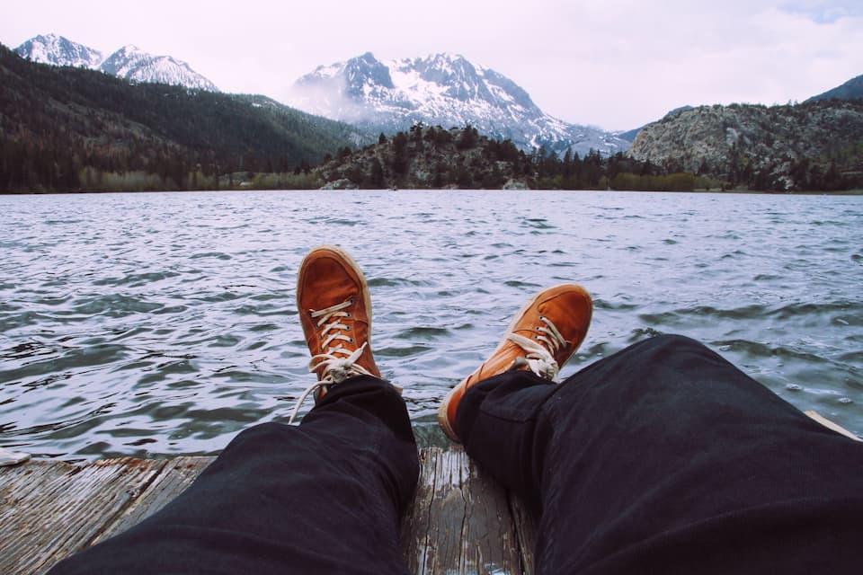 Pies descansando sobre un muelle de madera, con vista a un lago tranquilo y montañas al fondo.