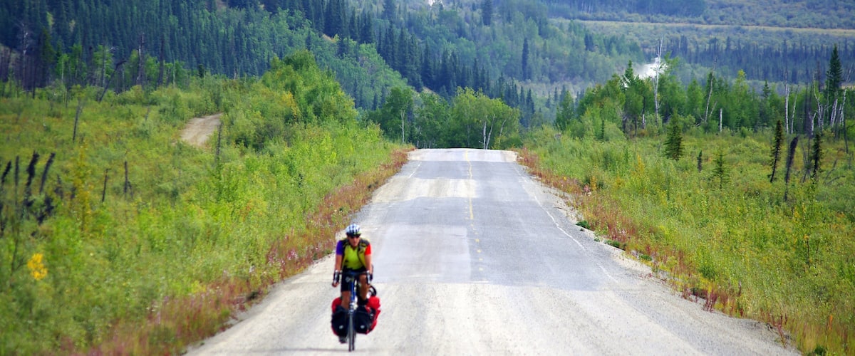 A cyclist on a gravel road surrounded by dense forest and a winding river in the distance.
