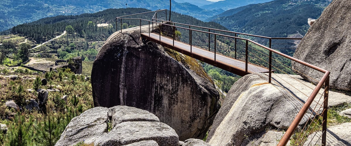 A wooden bridge with metal railings spanning a rocky gorge, overlooking a valley with mountains and trees.