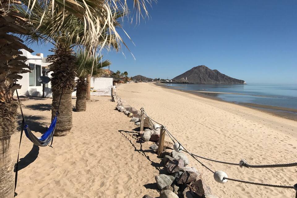 A sandy beach with palm trees, a hammock, and a rocky barrier.