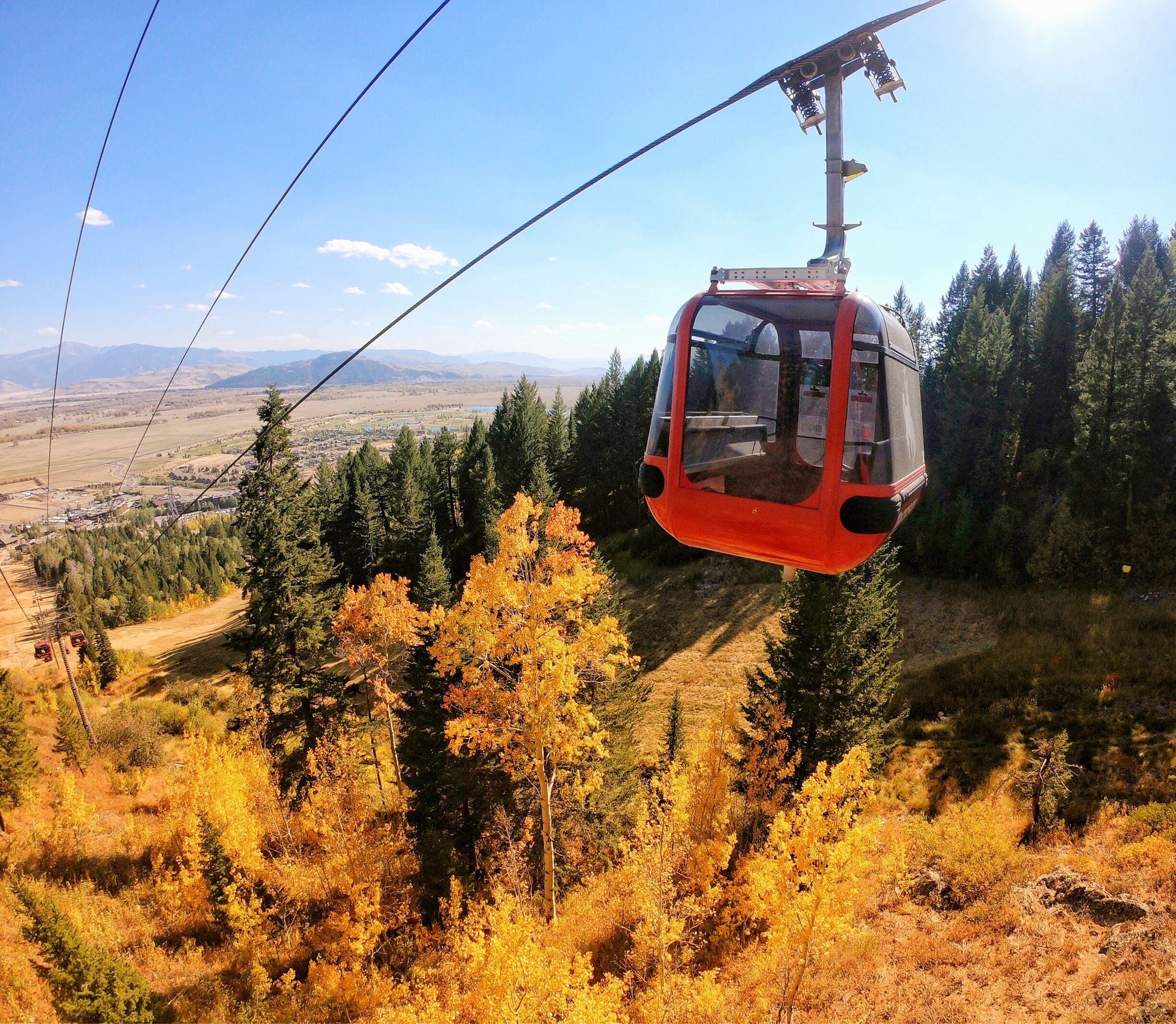 A red cable car suspended by cables, surrounded by autumn-colored trees and a distant view of a town and mountains.