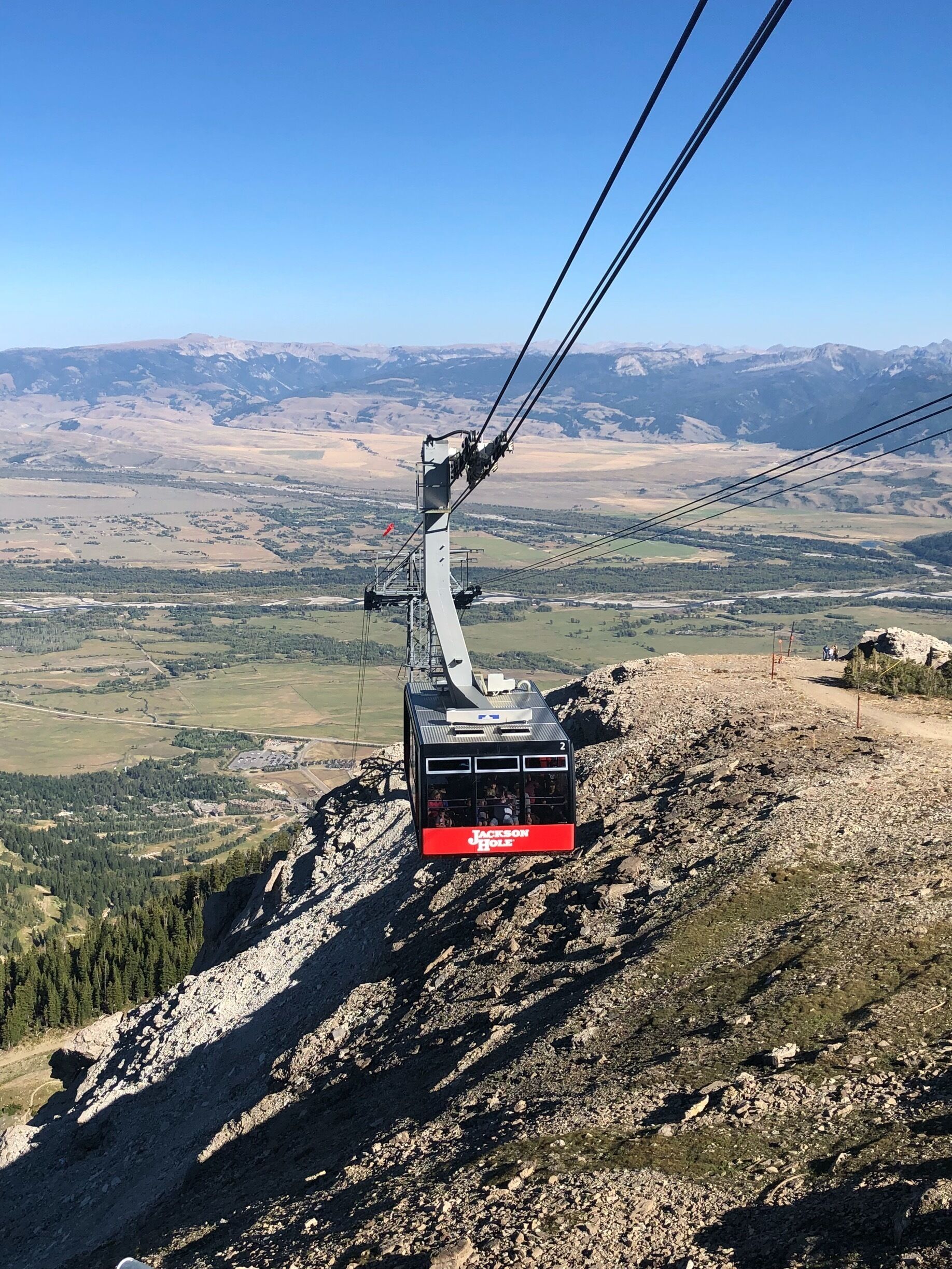 A cable car with 'Jackson Hole' written on it, suspended by cables, overlooking a mountainous landscape with valleys and forests.