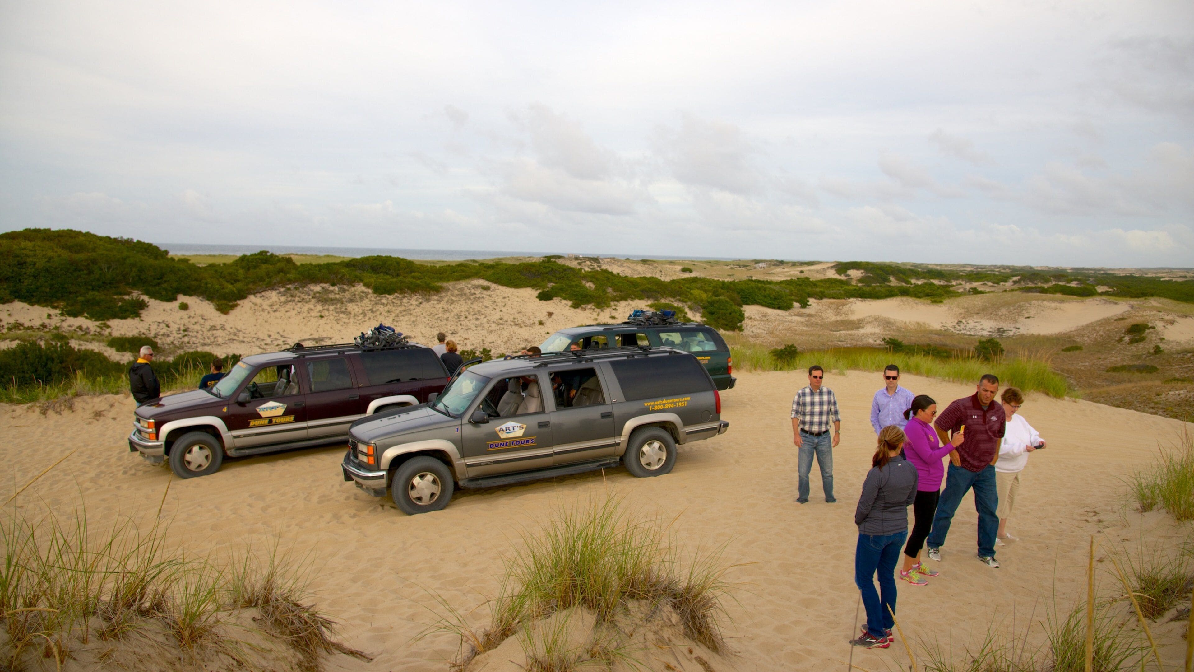 Vehículo turístico en un recorrido por dunas estacionado en una playa arenosa, con dunas al fondo.