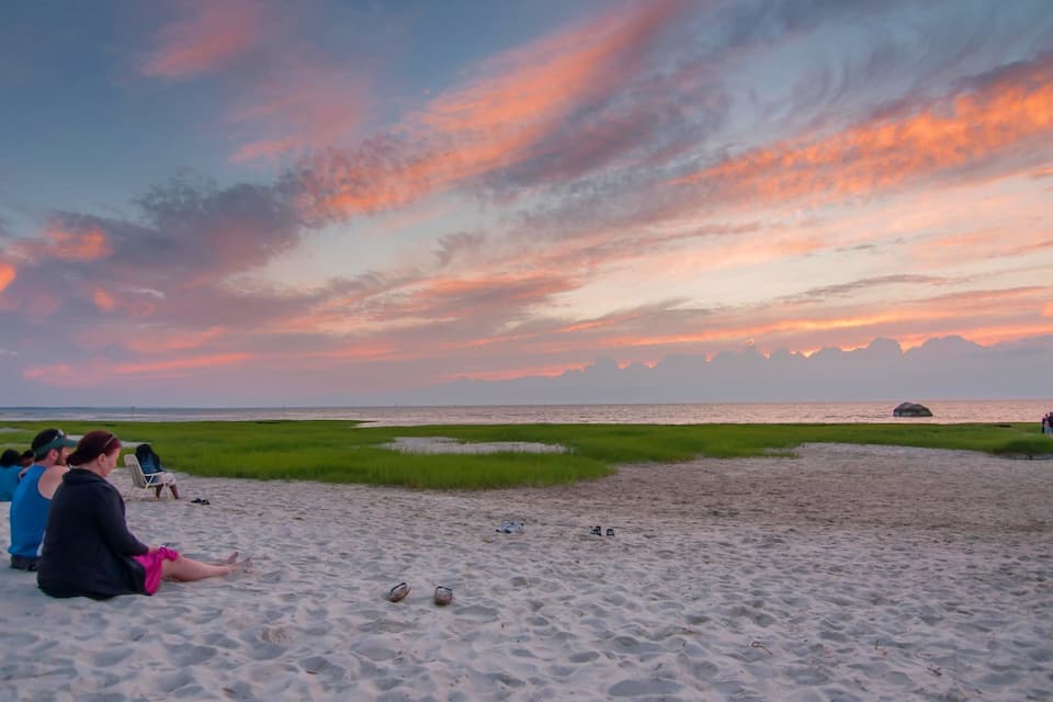 Una playa con gente sentada sobre la arena, un atardecer en el fondo y campos verdes a lo lejos.