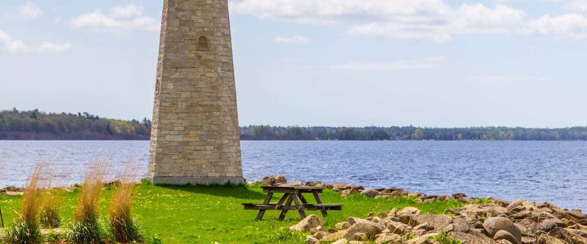 A stone lighthouse by a lake with a picnic table nearby.