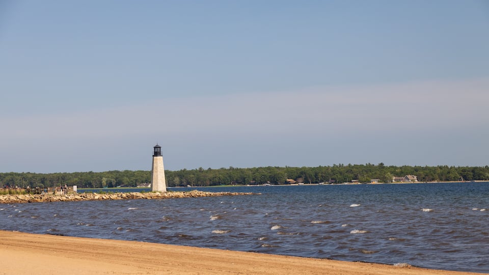 A lighthouse by a sandy beach with calm water and a forested shoreline.