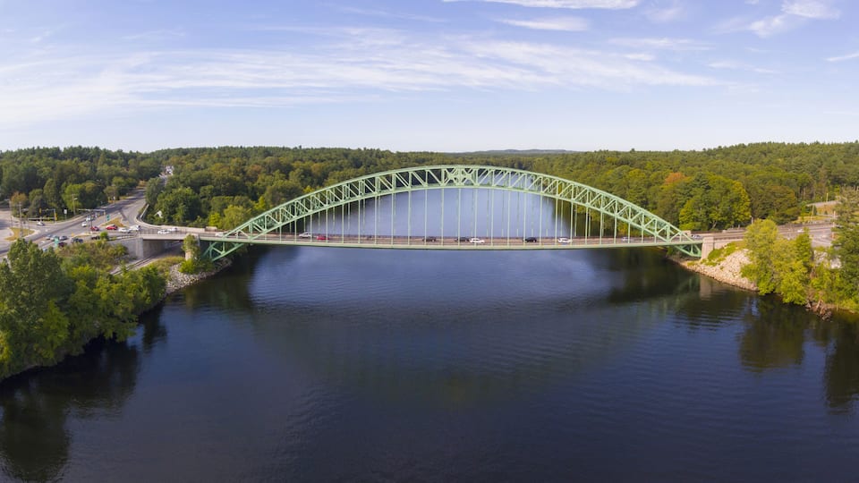 A green steel bridge over a calm river, surrounded by dense green forest and a clear sky.