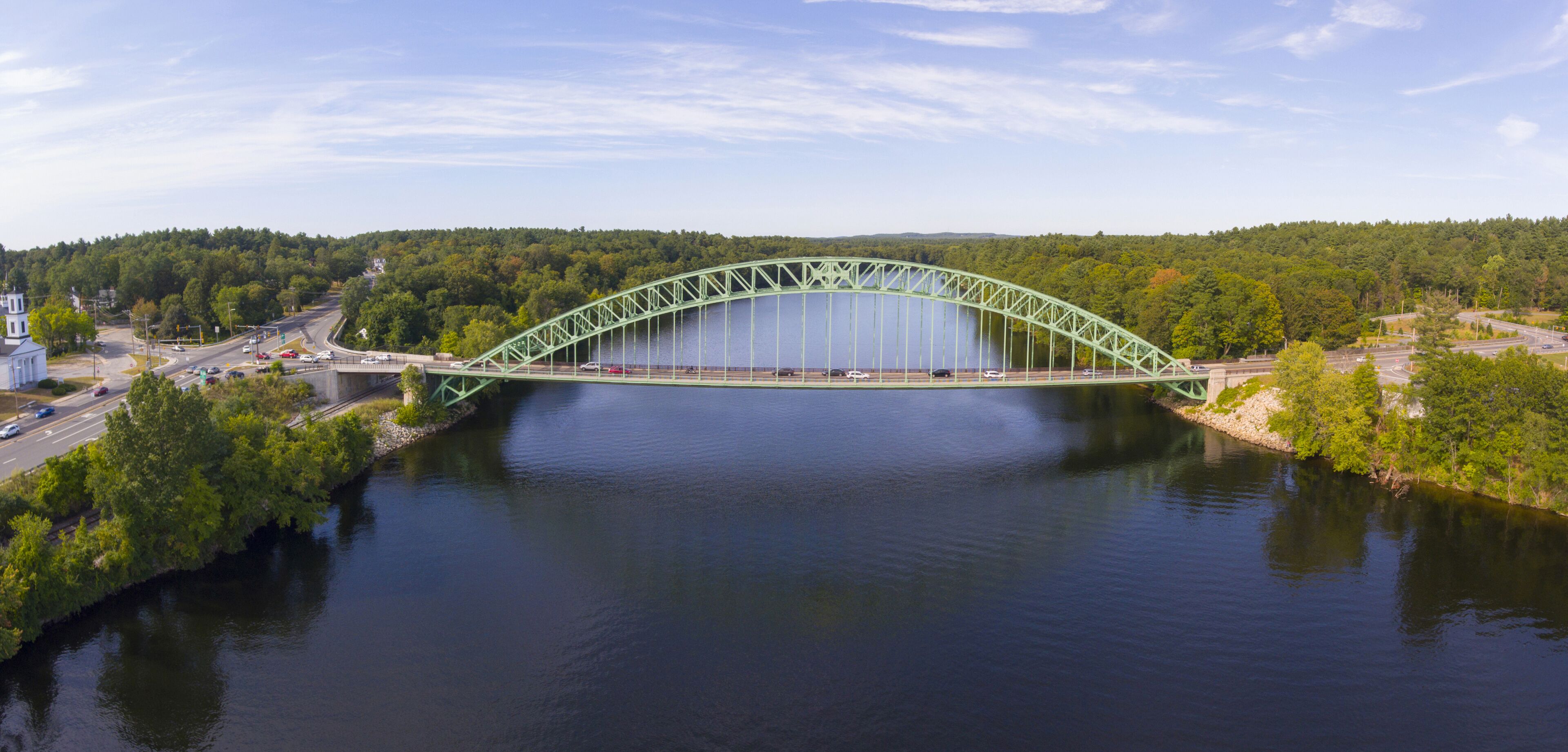 A green steel bridge over a calm river, surrounded by dense green forest and a clear sky.