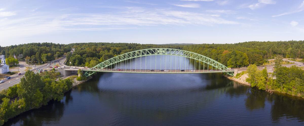 A green steel bridge over a calm river, surrounded by dense green forest and a clear sky.