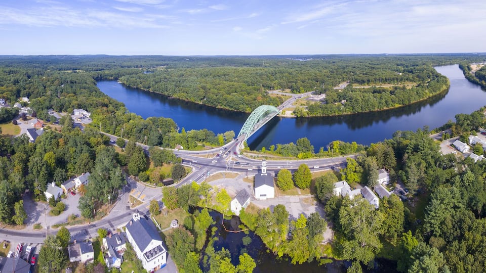An aerial view of a river with a bridge, surrounded by dense forests and residential areas.