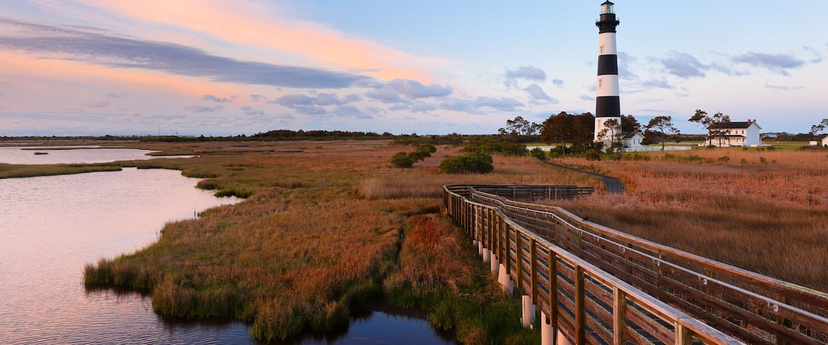 A lighthouse, a wooden bridge, and a marshy landscape with a sunset sky.