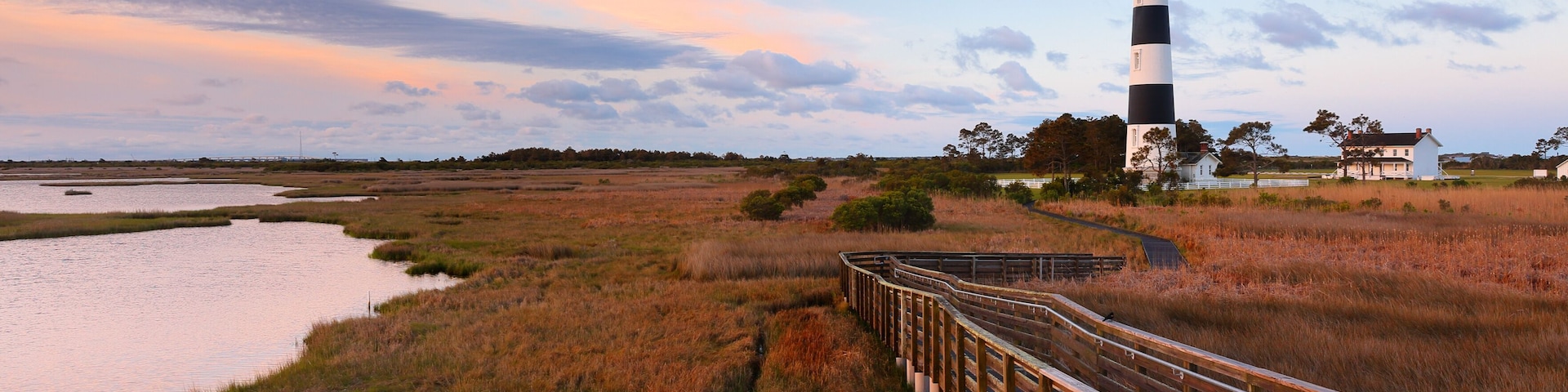 A lighthouse, a wooden bridge, and a marshy landscape with a sunset sky.