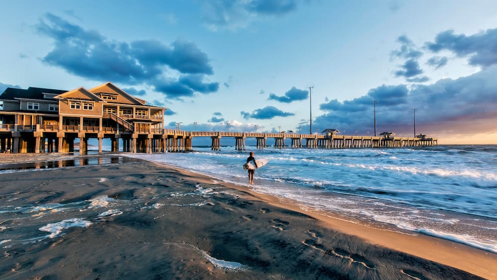 Une plage avec une jetée qui s’avance dans l’eau, une personne tenant une planche de surf et une grande maison construite sur pilotis.