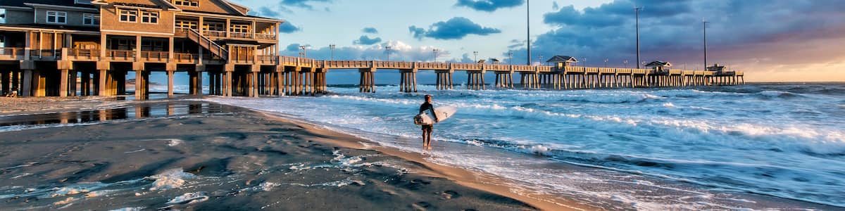 A beach with a pier extending into the water, a person with a surfboard, and a large house on stilts.