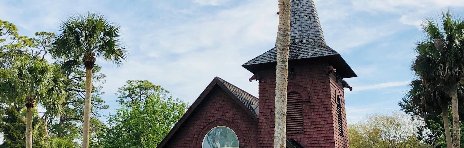 Une église dotée d’un clocher imposant et d’une façade en briques rouges, entourée d’une végétation luxuriante et de palmiers.