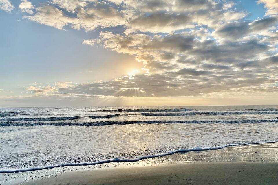 Una playa con olas, cielo nublado y el sol poniéndose o saliendo.