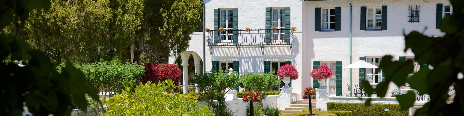 Une maison blanche aux volets verts et au toit recouvert de tuiles rouges, entourée de jardins et d’arbres bien entretenus.
