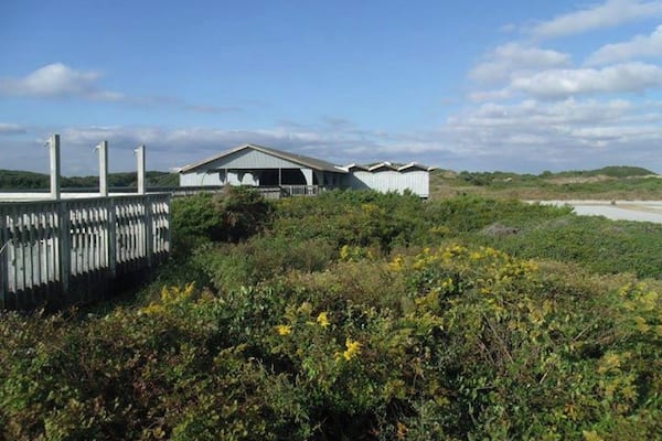 En kystbygning med hvidt tag og træterrasse, omgivet af frodig vegetation og en klar himmel.
