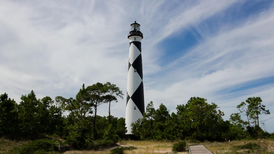 A lighthouse with a black and white striped pattern, surrounded by trees and a wooden boardwalk.
