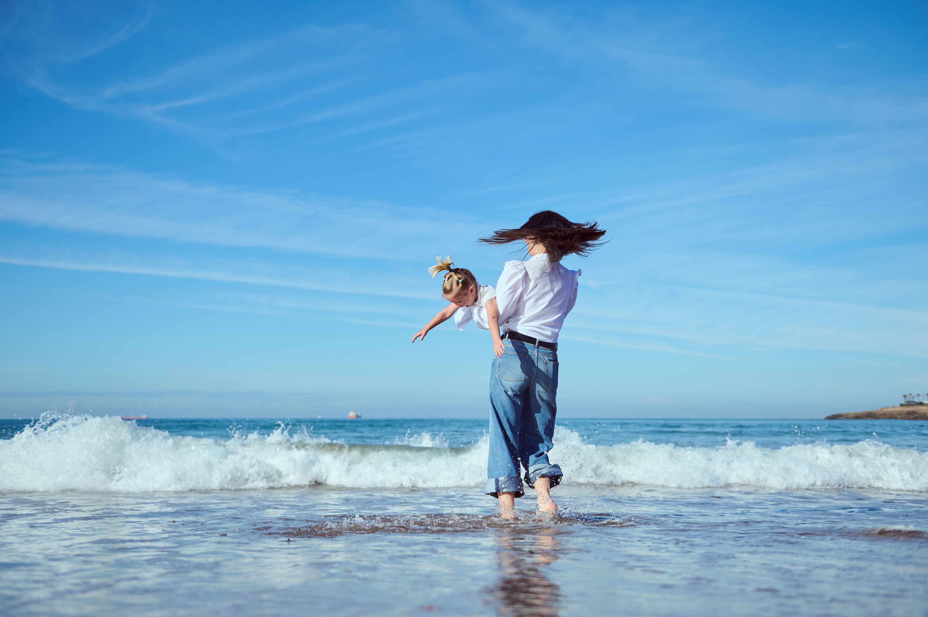 Cheerful young mom playing with her little daughter, running barefoot on the Atlantic beach, jumping on splashing waves