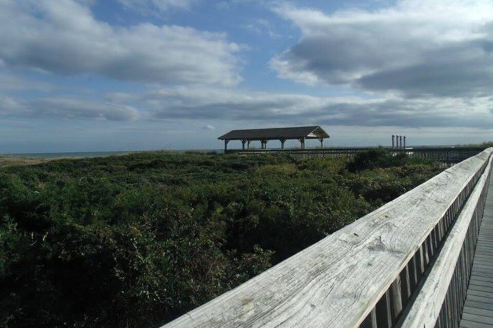 A wooden boardwalk leading to a covered viewing platform with a view of the ocean and industrial structures in the distance.