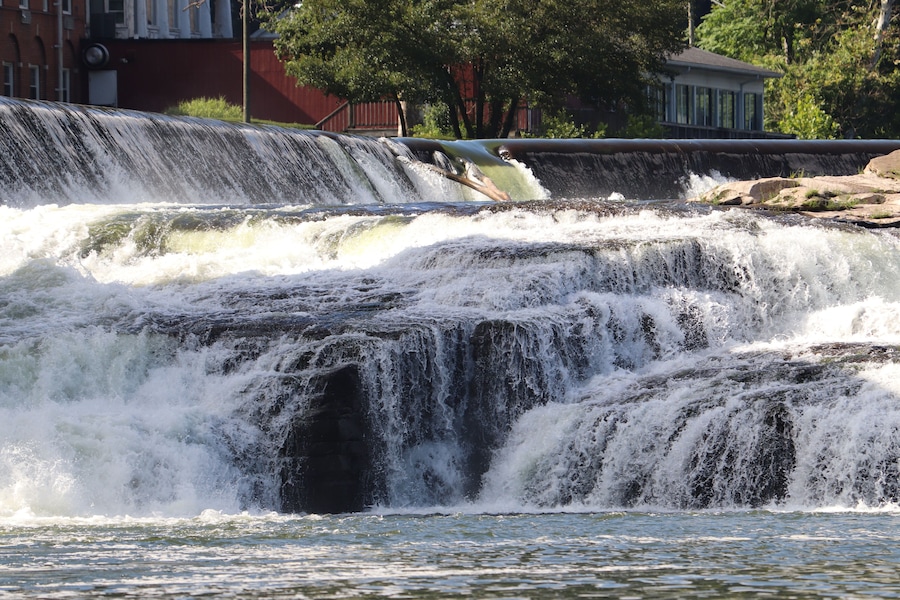 A multi-tiered waterfall cascading over rocks, surrounded by a cityscape with buildings and trees.
