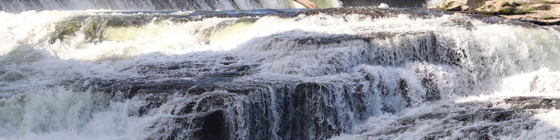 Uma cascata em vários níveis despencando sobre rochas, emoldurada por uma paisagem urbana com edifícios e árvores.
