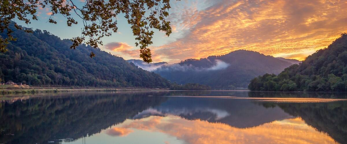 A calm lake with a wooden dock, surrounded by forested hills and a sunset sky.