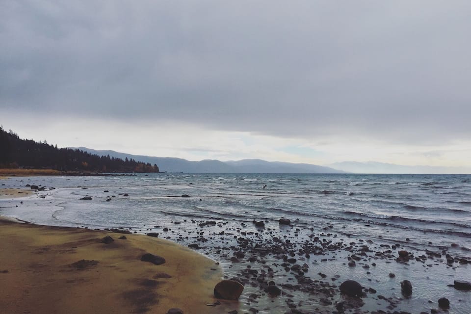 A rocky beach with waves, a forested hillside, and distant mountains under a cloudy sky.