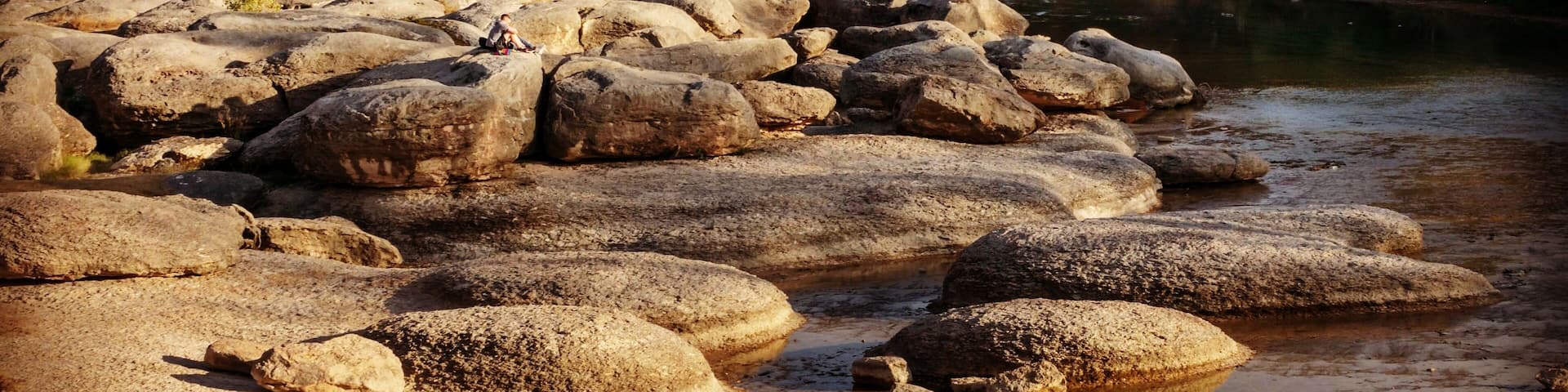 A rocky riverbank with large boulders and a forested hillside in the background.