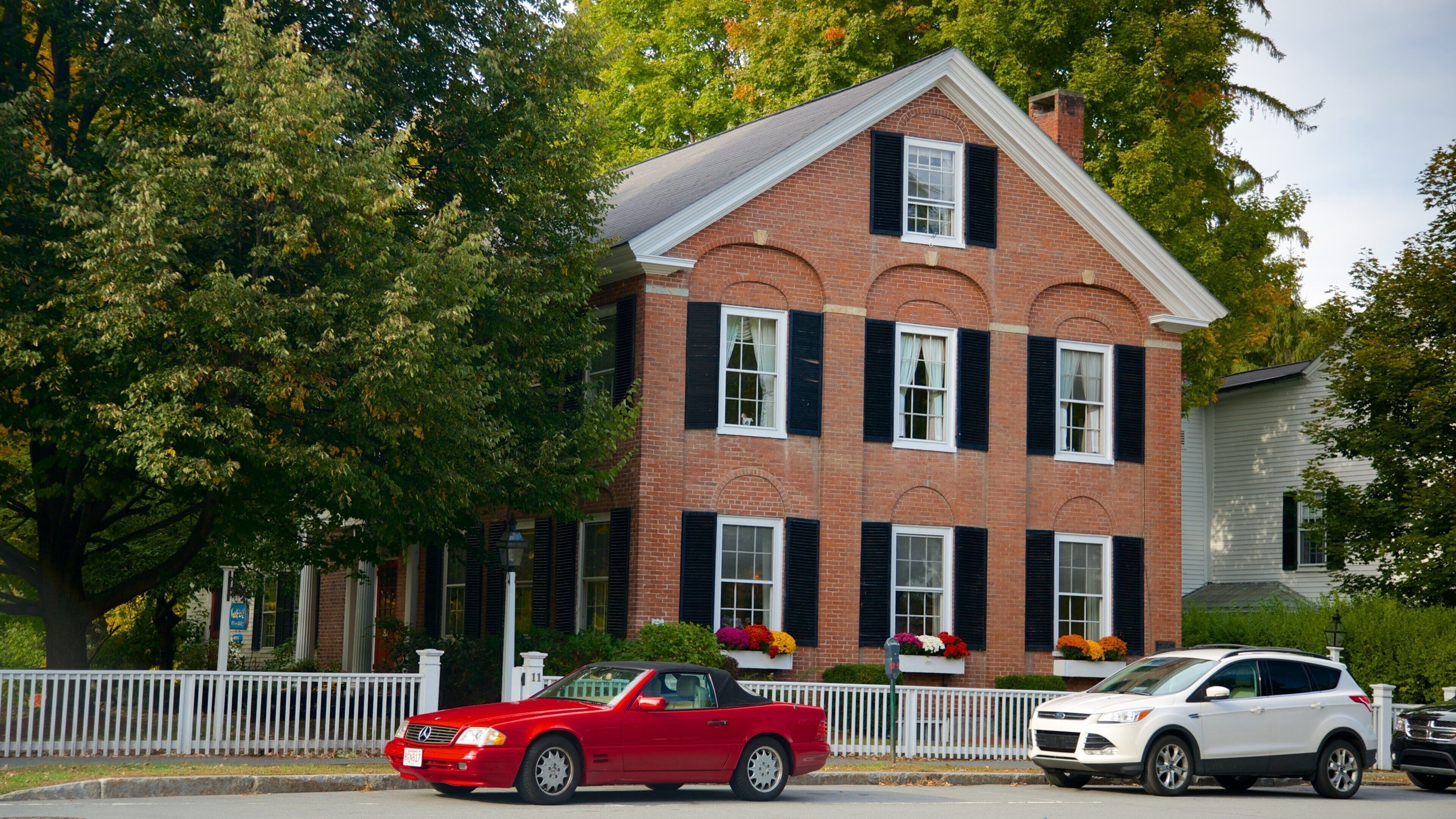 A red convertible car parked in front of a brick house with black shutters and a white picket fence.