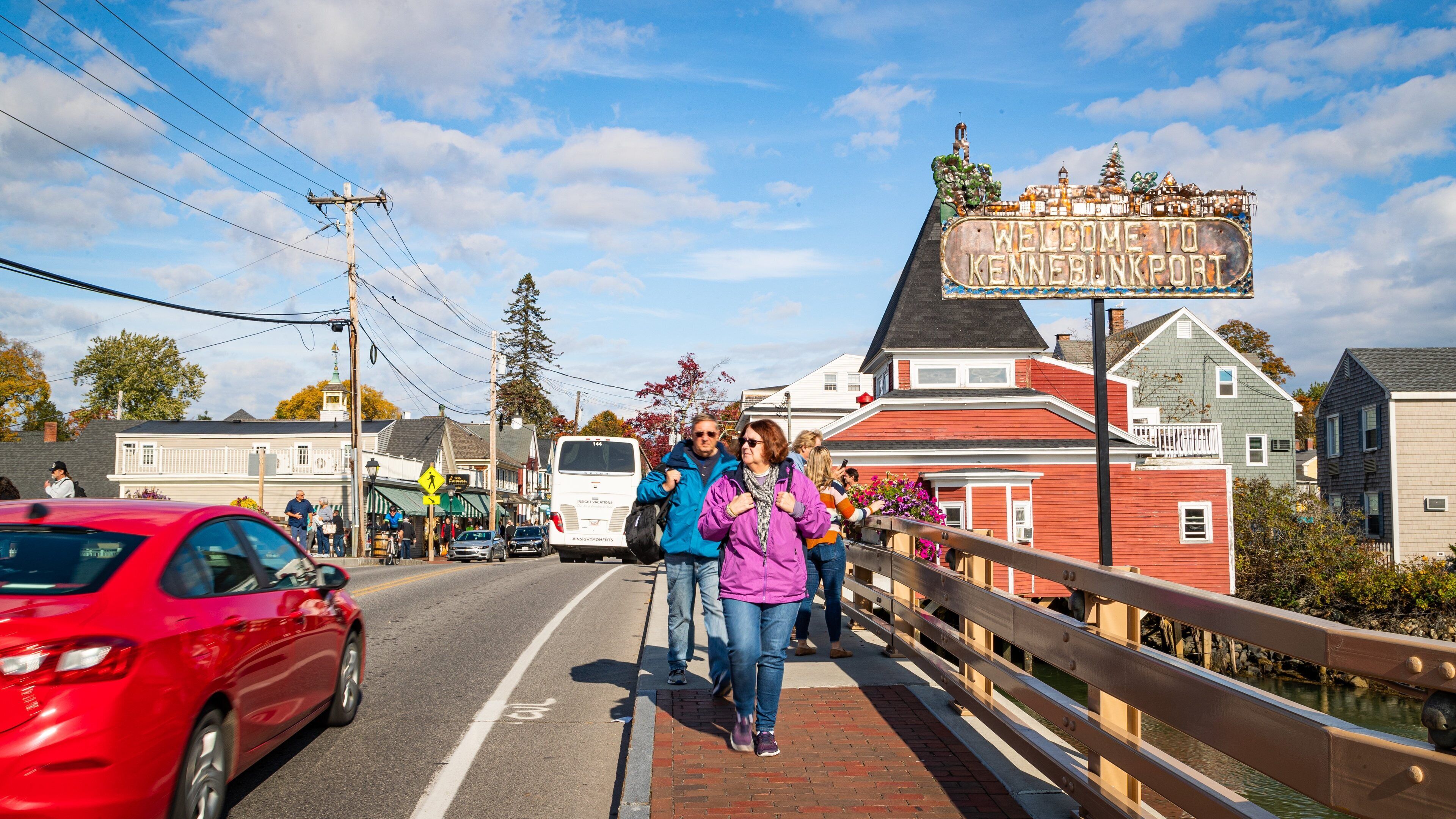 A pedestrian bridge with a 'Welcome to Kennebecport' sign, a red car, and a white van on the road.