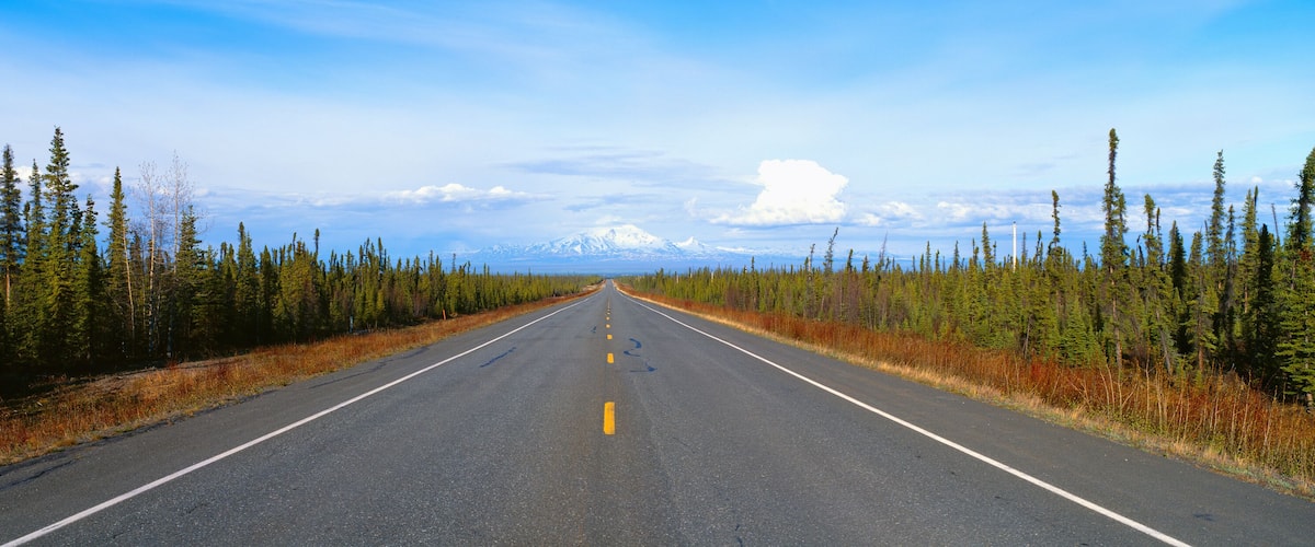 A straight road flanked by dense coniferous forests, leading towards distant snow-capped mountains under a clear sky.