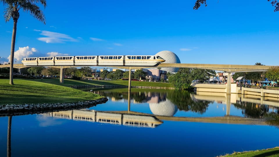 A train crossing a bridge over a calm body of water with a spherical building in the background.