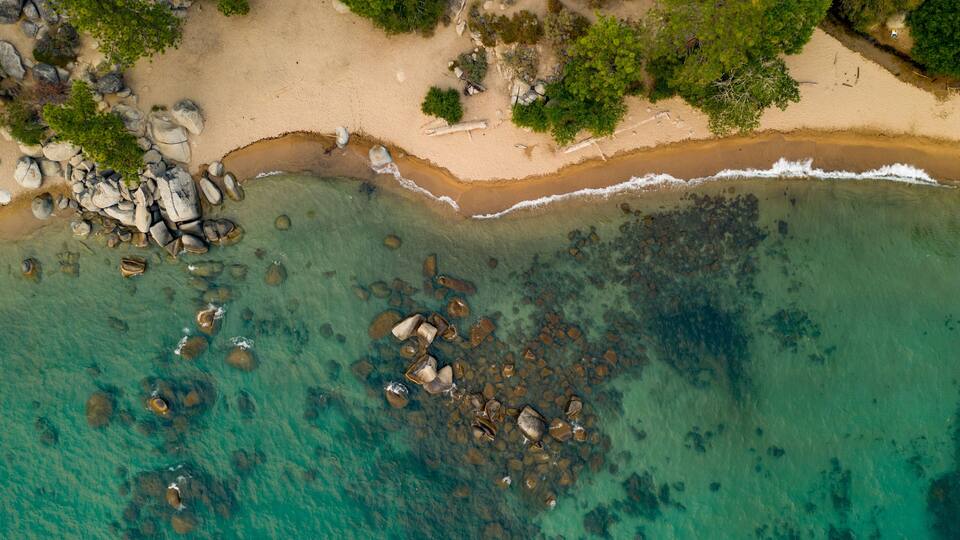 An aerial view of a beach with clear turquoise water, scattered rocks, and dense green foliage.