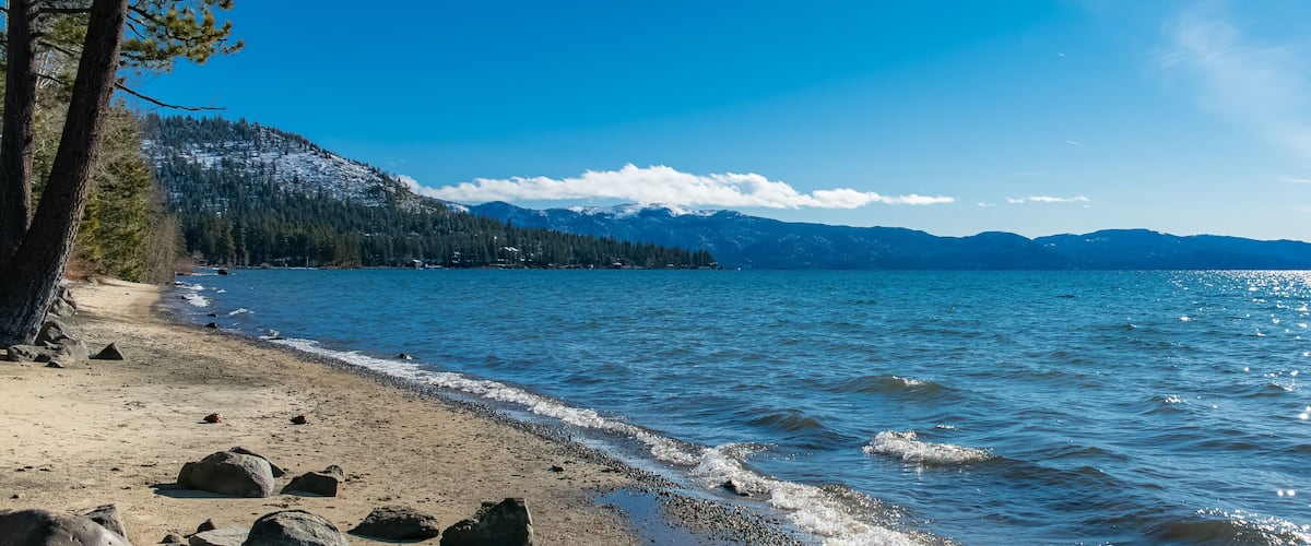 A clear lake with a sandy beach, surrounded by mountains and trees.