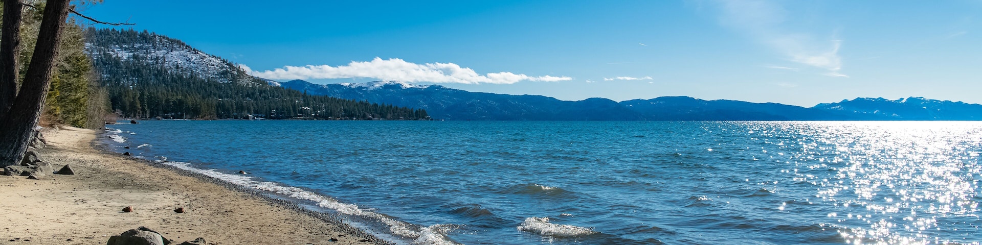 A clear lake with a sandy beach, surrounded by mountains and trees.