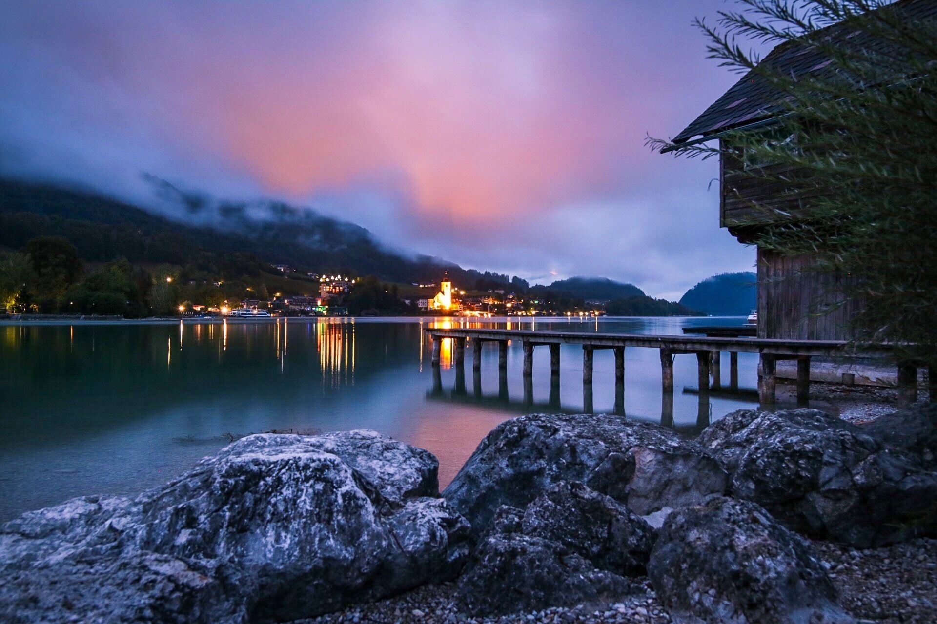 A calm lake at dusk with a wooden pier extending into the water, a small building on the right, and a lit-up church in the background.