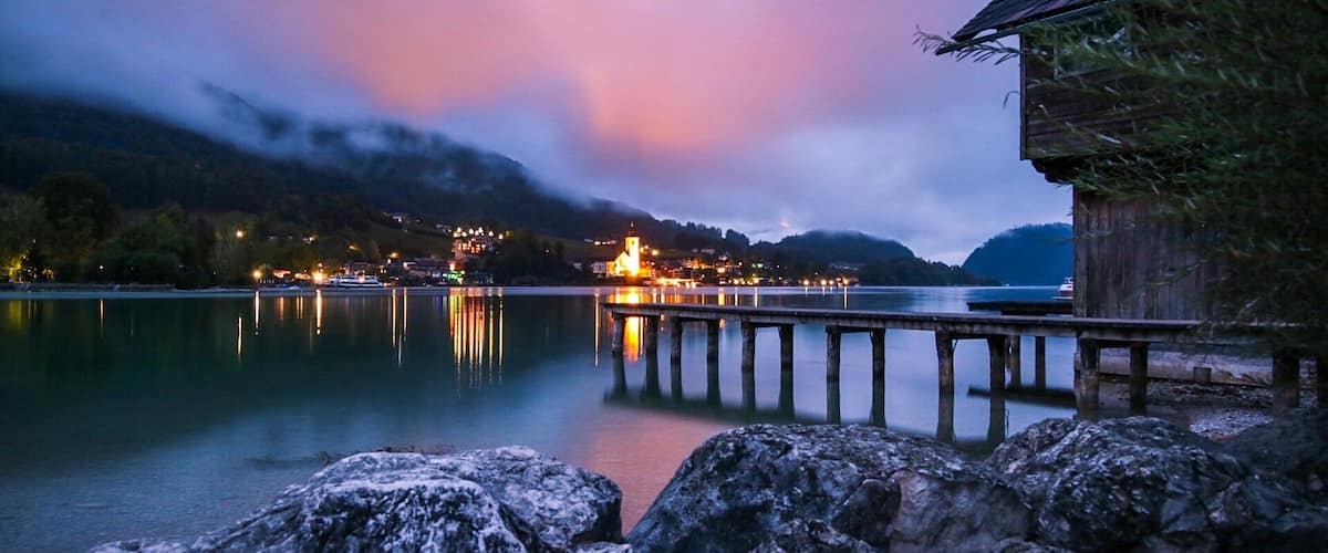 A calm lake at dusk with a wooden pier extending into the water, a small building on the right, and a lit-up church in the background.