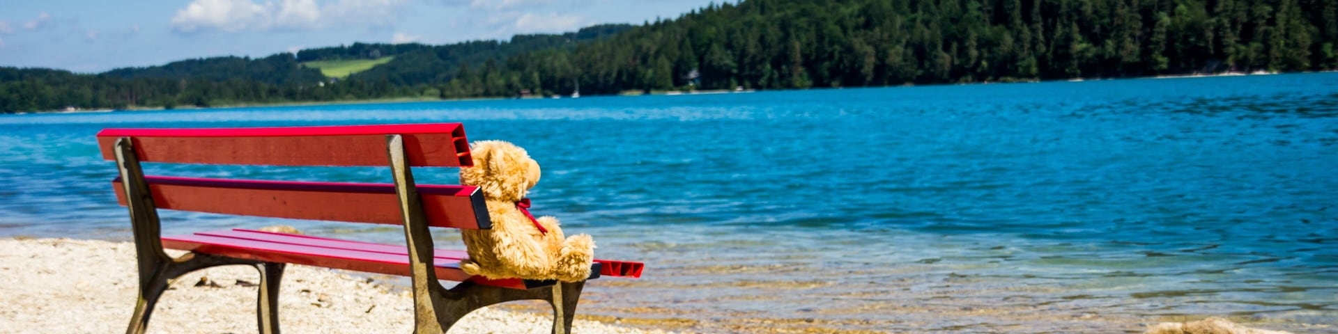 A red bench with a teddy bear on it, a pebble beach, and a lake with a forested hill in the background.