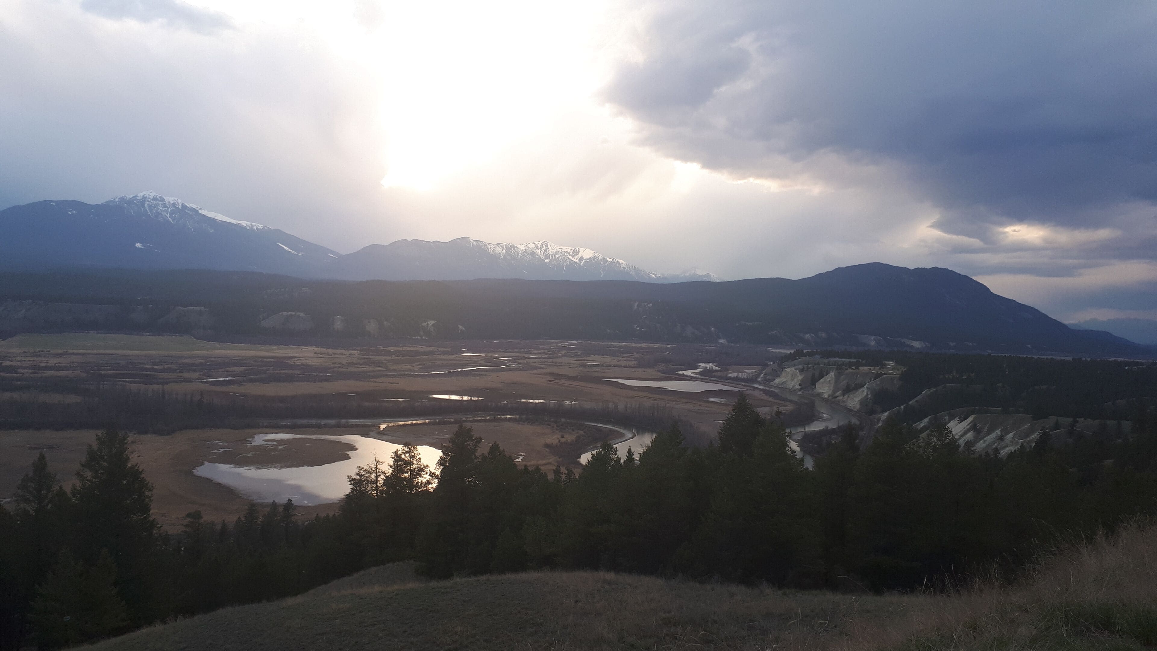 A mountainous landscape with snow-capped peaks, a river, and a forested foreground.