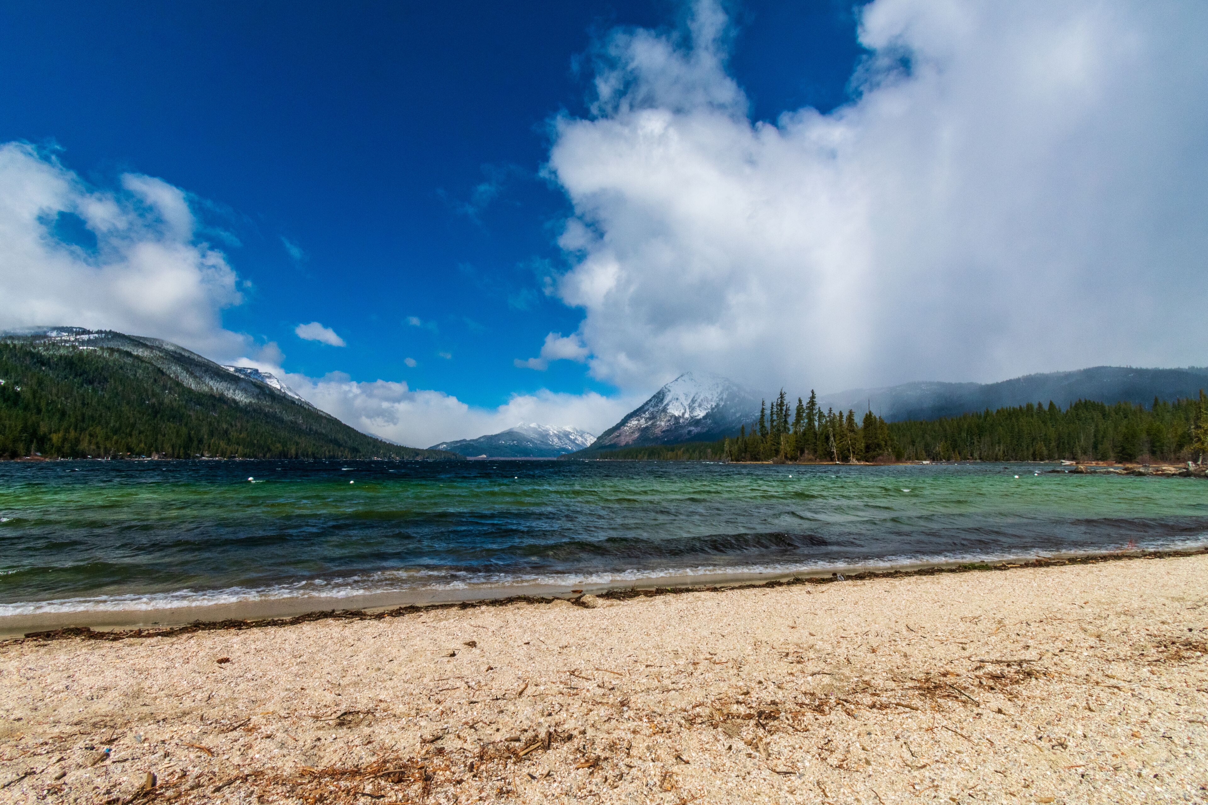 A lake with mountains in the background, a clear sky, and a sandy beach.