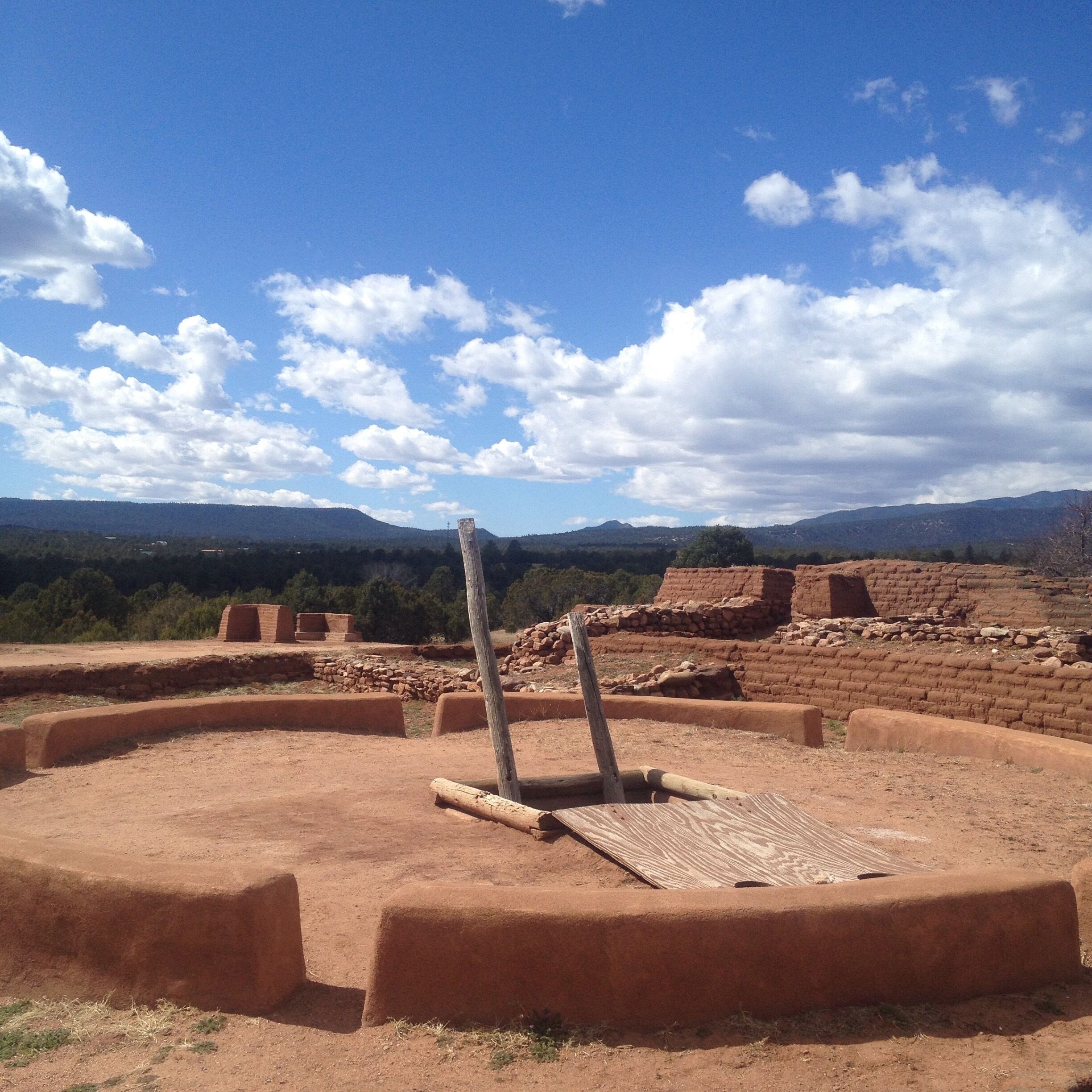 Ancient ruins with a circular stone structure and a wooden post in the center, surrounded by a mountainous landscape.
