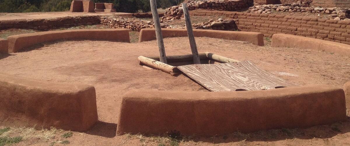 Ancient ruins with a circular stone structure and a wooden post in the center, surrounded by a mountainous landscape.