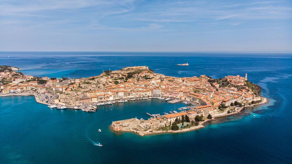 An aerial view of a coastal town with a fortified harbor, historic buildings, and a prominent lighthouse.