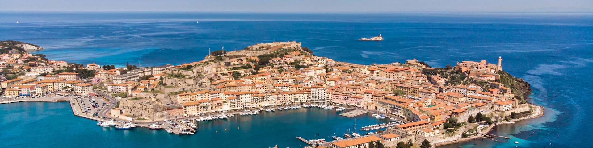 Vista aérea de un pueblo costero con un puerto fortificado, edificios históricos y un faro destacado.